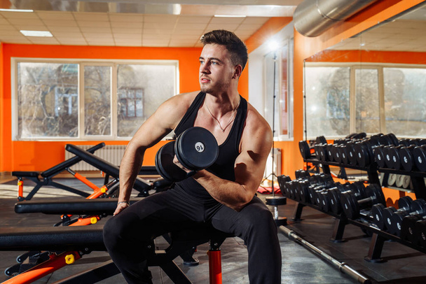 Portrait of athletic, handsome, young, muscular european brunette doing physical exercises, biceps workouts with dumbbells in gym, copy space. Wearing black t-shirt and pants. Looking and his muscles. - Photo, Image
