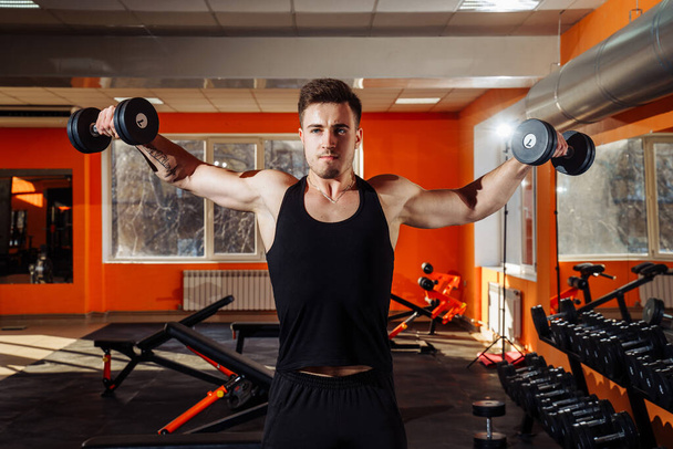 Athletic young man exercising his shoulders and doing side raises with pair of dumbbells at gym. Inflated, athletic, attractive guy pumping-up his hands hard works on his body - Photo, Image