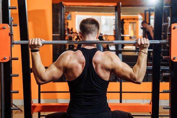 Average plan, back view. A muscular man dressed in black sportswear is training muscles, biceps and triceps - with an effort he lifting the bar. Fitness and sport. Healthy lifestyle concept - Photo, Image