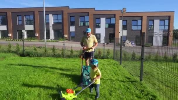 Der kleine Sohn mäht Gras mit dem Rasenmäher wie sein Vater. Gimbal bewegt sich nach vorne - Filmmaterial, Video