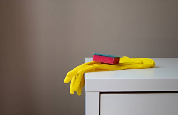 Yellow rubber gloves and a red sponge lie on the edge of a white table with copy space - Foto, Imagen