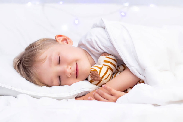 Charming little white boy sleeps in bed. hugs a soft toy - Fotoğraf, Görsel