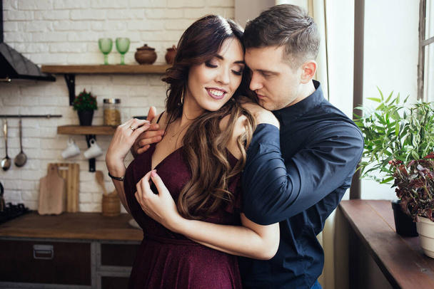 A lovely couple are standing in the kitchen with closed eyes. A smiling woman in dress with a yellow tulips in her hand. Harmonious family. Romantic love story. Happiness.  - Фото, зображення