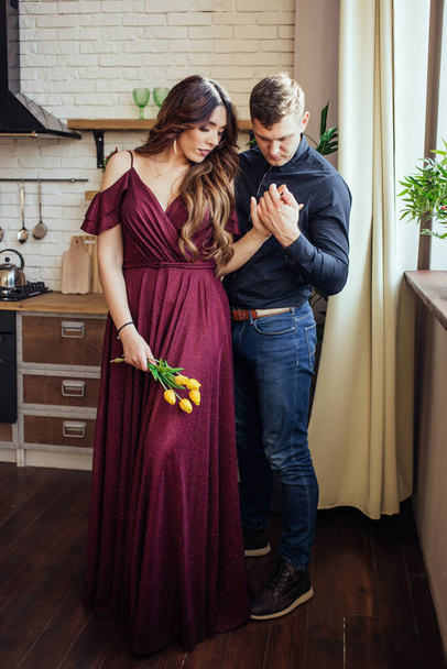 A lovely couple are standing in the kitchen. Her hand in his hand. A woman in dress with a yellow tulips in her hand. Harmonious family. Romantic love story. Happiness.  - Fotografie, Obrázek