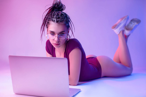 das schlanke Mädchen auf weißem Hintergrund in Neonfarbe liegt auf dem Boden und blickt auf einen Laptop. Übungen und Training online lernen - Foto, Bild
