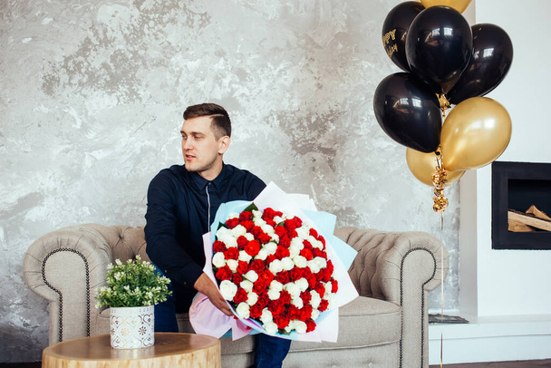 Een man in een blauw shirt met een groot boeket rode en witte rozen zit thuis op de bank. Gelukkige verjaardag. Zwarte en gouden verjaardagsballonnen. Thuis comfort. Wachten op een vrouw. Levensstijl.  - Foto, afbeelding