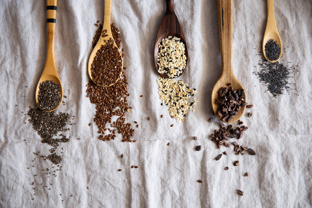 Nasiona szałwii, nasiona lnu, sezam, szczypce kakaowe i nasiona maku na drewnianych łyżeczkach na rustykalnym materiale. - Zdjęcie, obraz