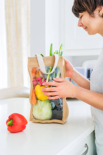 Woman holding reusable eco sackcloth fabric bag packaging with fresh organic vegetables on white kitchen table. Home delivery concept. Local farmer healthy food. Zero waste. Vertical card. Copy space - Photo, Image