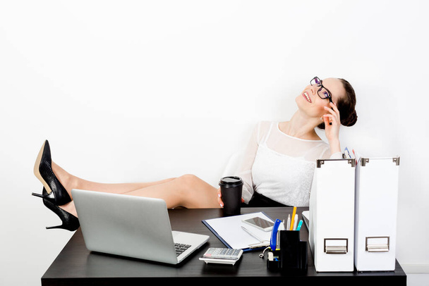 A young handsome business woman in strict clothes and eyeglasses cozily threw feet in shoes the desk and closed her eyes dreaming with a smile on her face on a white background in front of her laptop. The concept of a comfortable workplace. - Foto, immagini