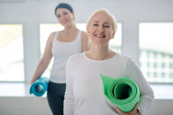 Blonde female standing with yoga rug and smiling, brunette female standing behind her - Фото, зображення