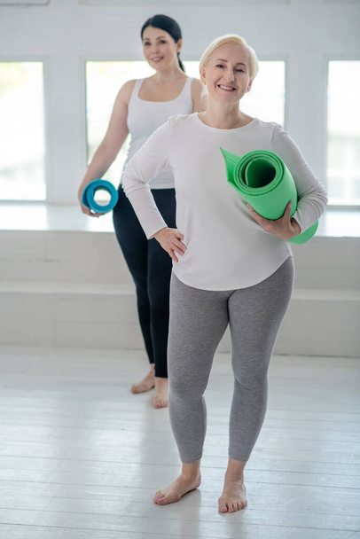 Blonde female holding yoga rug and smiling, her one hand on her waist, brunette female standing behind her - Foto, imagen