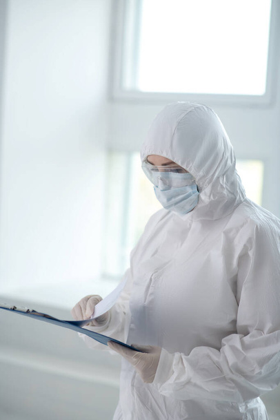 Medical worker in protective clothing, medical mask and goggles looking through papers on clip folder - Fotografie, Obrázek