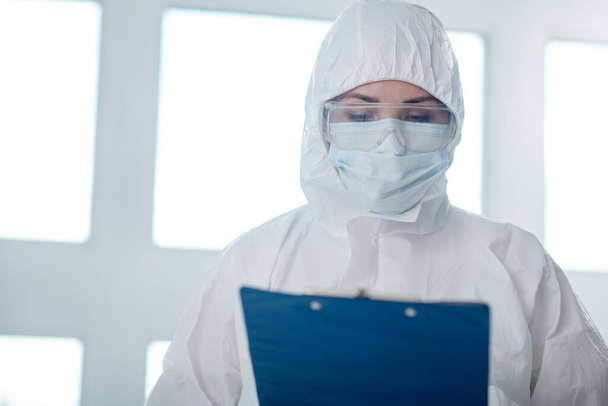 Medical worker in protective clothing, medical mask and goggles looking at clip folder - Foto, Imagen