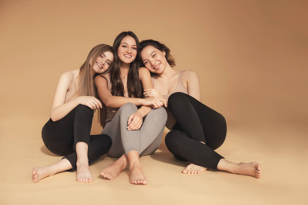 Glückliche junge multikulturelle asiatische Frauen, kaukasische Umarmungen und Lachen im Studio auf beigem Hintergrund. Beauty-Spa-Behandlung Hautpflege-Konzept - Foto, Bild