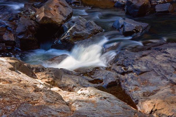 Agua intencionalmente borrosa por la lenta velocidad del obturador de la cámara que fluye suavemente sobre las rocas en un lecho de arroyo
 - Foto, imagen