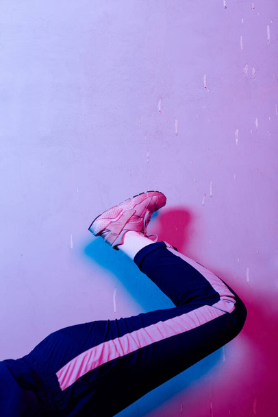 Crop female leg in blue sport pants and pink sneaker raised high in pink neon light - Foto, Bild