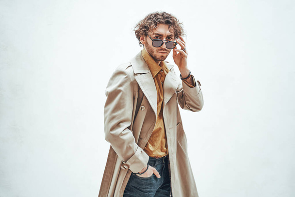 Στοχαστικός και όμορφος νεαρός που ποζάρει σε ένα φωτεινό στούντιο σε λευκό φόντο, κοιτώντας συγκεντρωμένος φορώντας καμπαρντίνα και γυαλιά, κοιτάζοντας κατευθείαν στην κάμερα - Φωτογραφία, εικόνα