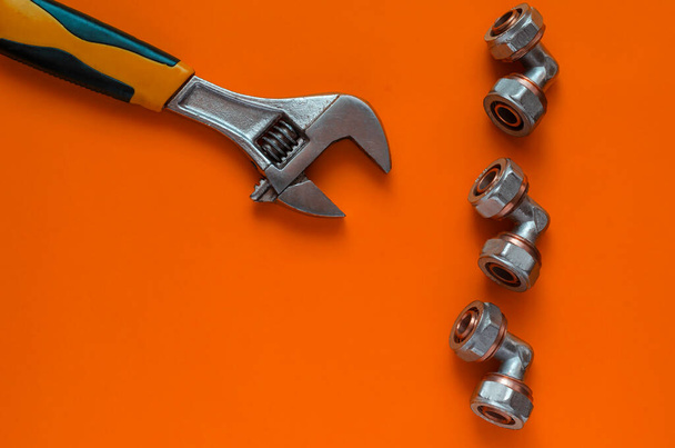 Creative orange background with hand tools and plumbing fittings. Adjustable wrench and three bronze corner fittings for multilayer water pipes. View from above. - Photo, Image