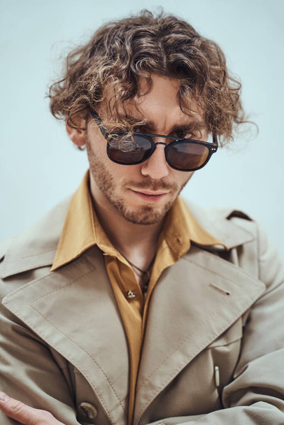 Jeune homme réfléchi et beau posant dans un studio lumineux sur un fond blanc, regardant concentré tout en portant un trench coat et des lunettes, regardant droit dans la caméra
 - Photo, image