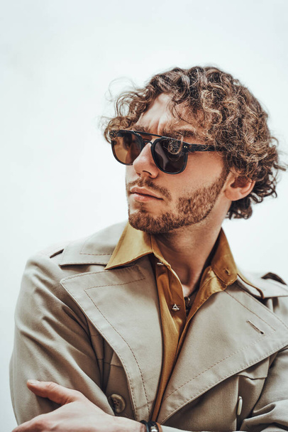 Cooles und gut aussehendes junges erwachsenes männliches Model, das in Trenchcoat, orangefarbenem Hemd und Sonnenbrille vor weißem Hintergrund für ein Fotoshooting posiert - Foto, Bild