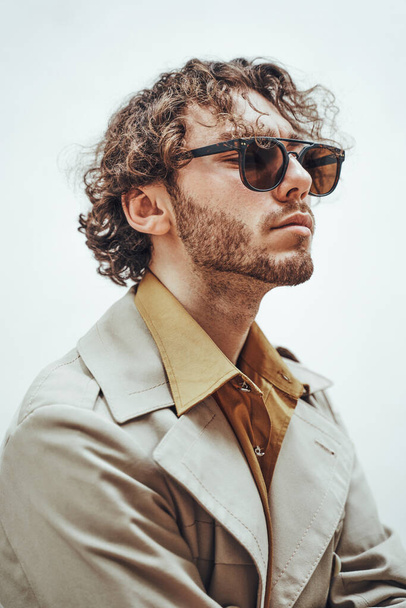 Detailní portrét elegantního mladého mužského modelu s kudrnatými vlasy pózující na bílém pozadí a nositelem trenčové bundy se slunečními brýlemi - Fotografie, Obrázek