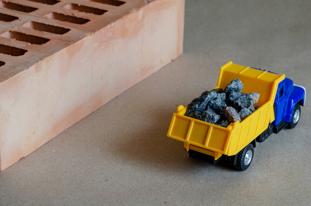 Ένα κίτρινο σκουπιδιάρικο μεταφέρει συντρίμμια δίπλα σε ένα τούβλο. Φορτηγό παιχνιδιών με πλήρες σώμα οικοδομικών υλικών. Δημιουργικό βιομηχανικό υπόβαθρο. Κατασκευή και μεταφορά οικοδομικών υλικών. - Φωτογραφία, εικόνα