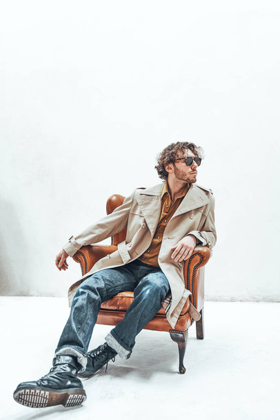 Schöner junger Kerl mit lockigem Haar posiert in einem hellen Studio, trägt beigen Trenchcoat und Sonnenbrille, sieht selbstbewusst und cool aus, während er auf einem Lederstuhl sitzt - Foto, Bild