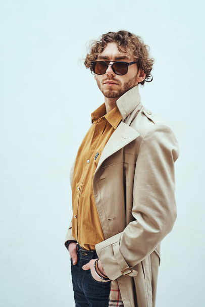 Елегантно виглядає молодий дорослий кавказький чоловік позує в яскравій студії на білому тлі, одягнений в траншею пальто і сонцезахисні окуляри, тримаючи руки в кишенях
 - Фото, зображення