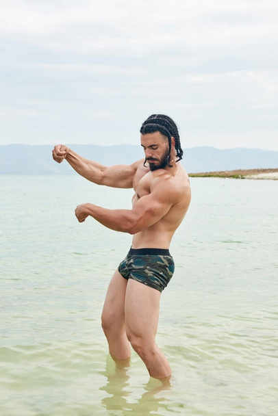 young man on the beach, young muscular man exercising on the beach, young muscular man doing bodibuilding exercises on the beach, athletic young man on the beach - Zdjęcie, obraz