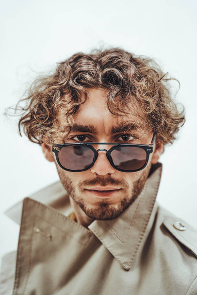 Nachdenklicher und gut aussehender junger Mann posiert in einem hellen Studio vor weißem Hintergrund, blickt konzentriert in Trenchcoat und Brille, direkt in die Kamera - Foto, Bild