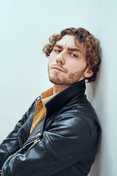 Quente e assertivo, jovem modelo masculino posando em um estúdio para a sessão de fotos vestindo casaco de couro elegante, olhando assertivo enquanto se inclina na parede
 - Foto, Imagem
