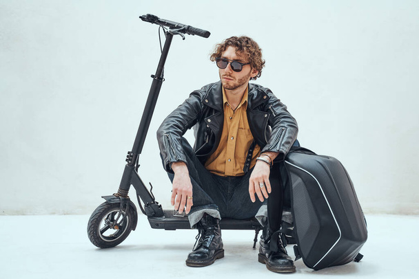 Stylish and self-assured, young male model posing in a studio for the photoshoot wearing fashionable leather jacket and sunglasses, looking cool and modern while relaxing on his scooter - Φωτογραφία, εικόνα