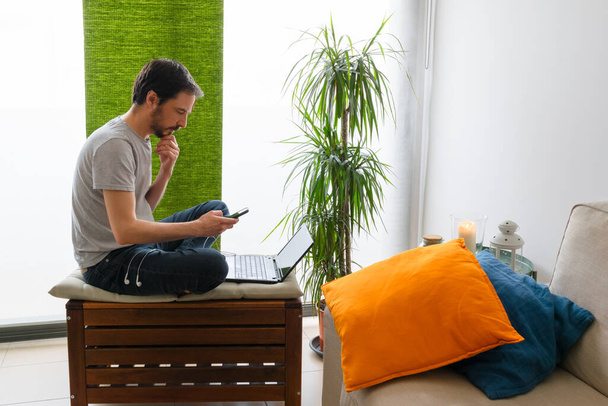 Hombre mirando el teléfono inteligente sentado descalzo en taburete de madera con cojines de colores
 - Foto, Imagen