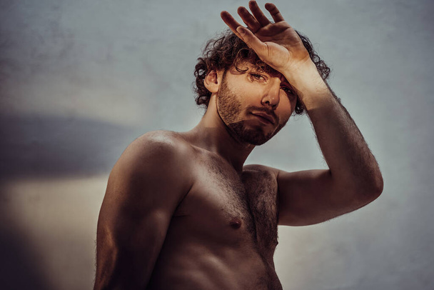 Homme nu et attrayant aux cheveux bouclés posant pour une séance photo sur un fond gris se couvrant de lumière avec une main
 - Photo, image