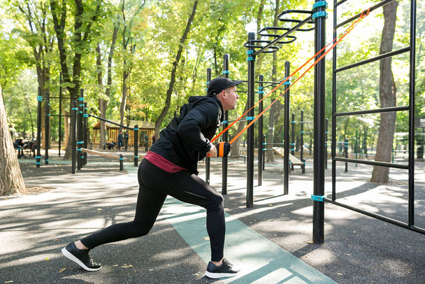 Vista lateral del hombre en ropa deportiva negra haciendo ejercicio con banda elástica en el campo de deportes del parque
 - Foto, Imagen