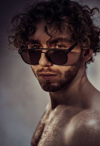 Heißer und glatter junger kaukasischer Mann mit lockigem Haar und Sonnenbrille posiert in einem dunklen Studio mit grauem Hintergrund und sieht sexy aus, während er seinen Körper zeigt - Foto, Bild