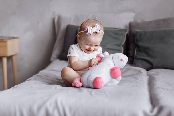 adorable little girl is playing with toy unicorn on bed at home. concept of childhood day. happy baby's, family day - Photo, Image