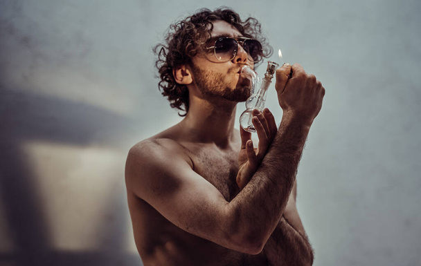 Соблазнительный и привлекательный молодой мужчина-модель с вьющимися волосами и солнечными очками позирует, будучи обнаженным и освещенным мягким солнечным светом в студии во время курения от крошечного стеклянного бонга
 - Фото, изображение