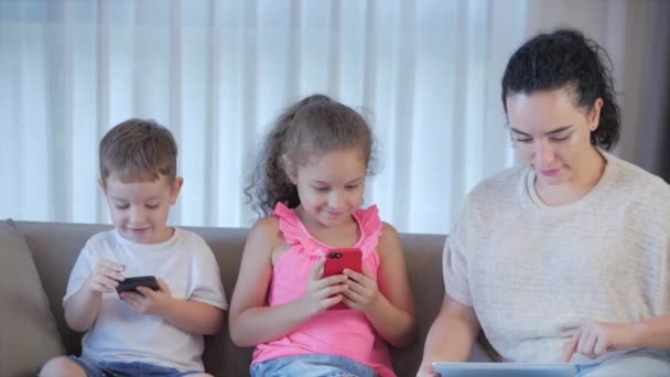 Happy family,mom and cute little kids,mother playing with children at home relaxing use a smartphone cuddling sit on sofa daughter and son laugh,watch funny videos,have fun,enjoy family life moments - Πλάνα, βίντεο