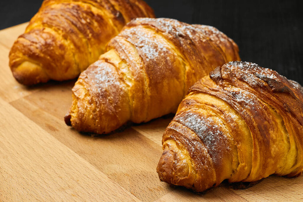 Three croissants with sugar powder on a wooden desk on a black background. Homemade baking concept. - Photo, Image