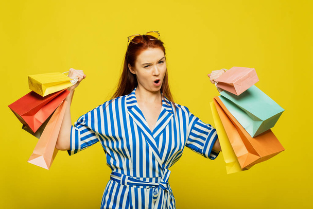 Pretty woman on a yellow background rejoices to bags with clothes after shopping. The girl shows strength and how many purchases she made. - Photo, Image