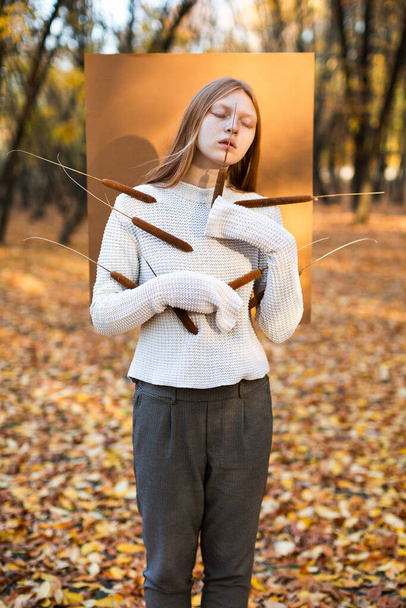 Sensual woman demonstrating comfortable clothes in autumn colors holding few cattails against carton in woods - Фото, изображение
