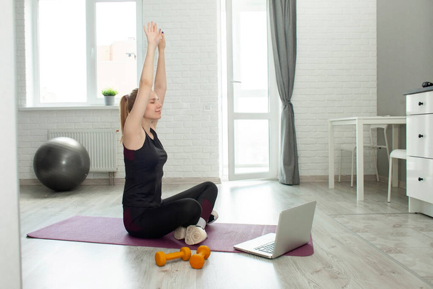 Genç sporcu kız evde dizüstü bilgisayarla yoga yapıyor, bir atlet odada Lotus pozisyonunda meditasyon yapıyor ve dizüstü bilgisayardan bir video izliyor, genel plan - Fotoğraf, Görsel