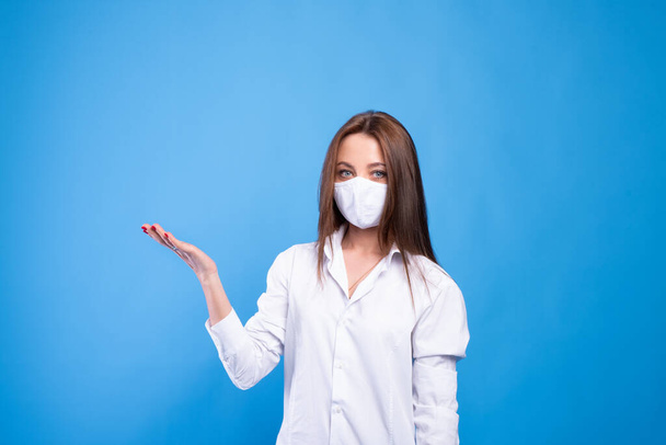 Beautiful caucasian young woman in white t-shirt with disposable face mask. Protection versus viruses and infection. Studio portrait, concept with blue background. Holds hand with palm up - Fotografie, Obrázek