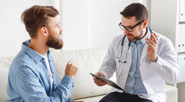 Young bearded guy gesticulating and talking with friendly male doctor while sitting on sofa in office of modern hospita - Photo, Image