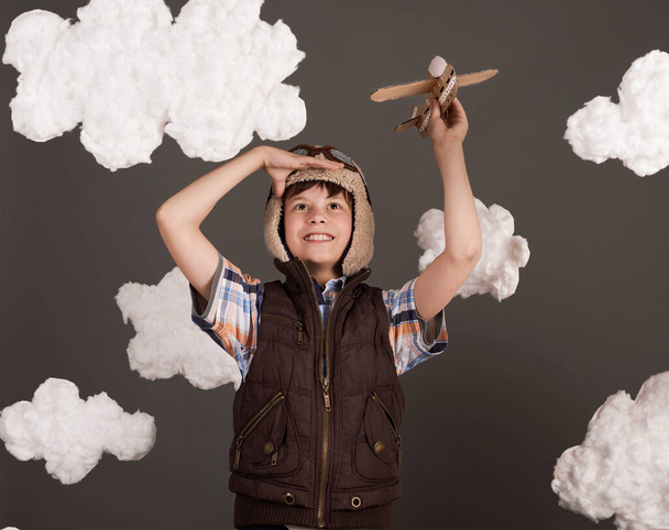 a boy plays with a cardboard airplane and dreams of becoming a pilot, dressed in a retro style jacket and helmet with glasses, clouds of cotton wool, gray background, tinted in brown - Photo, Image