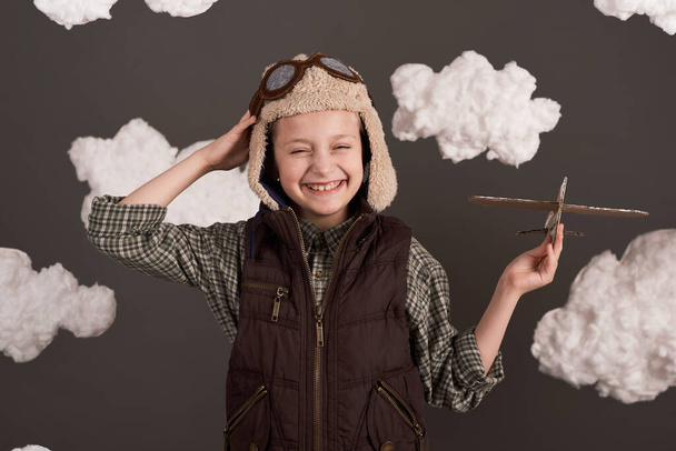 een meisje speelt met een kartonnen vliegtuig en droomt ervan piloot te worden, gekleed in een retro jasje en helm met bril, wollen wolken, grijze achtergrond, bruin getint - Foto, afbeelding