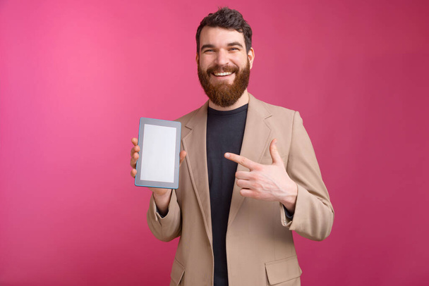Sonriente hombre barbudo está apuntando a su tableta mientras está de pie sobre fondo rosa
. - Foto, Imagen