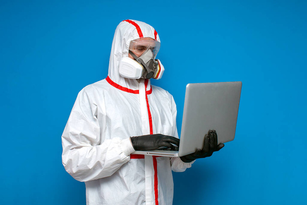 desinfiointityöntekijä suojapuvussa ja hengityssuojain tarkistaa koronavirustilastot kannettavalla tietokoneella, lääkäri virologi sinisellä taustalla
 - Valokuva, kuva