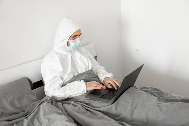 Man in protective white suit and medical mask is working from home in a bed with laptop because of coronavirus epidemic. Remote work during pandemic. Stay home during COVID-19 quarantine concept. - Fotó, kép